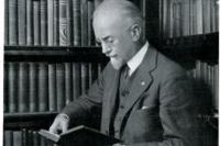 An old white man who is standing up and reading a book at a library